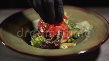 双手戴黑色食品手套，将切碎的西红柿放在盘子上，放在桌面上，隔离在黑色背景上。 食物